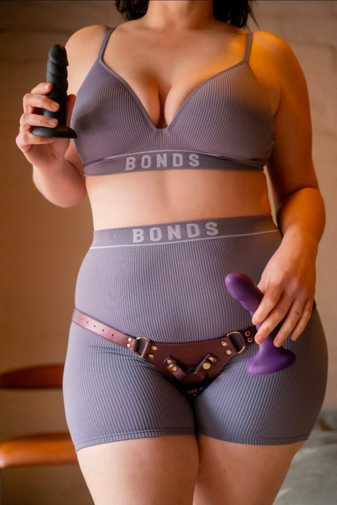 Ember wearing a strap-on and holding up two dildos, ready to start a pegging session